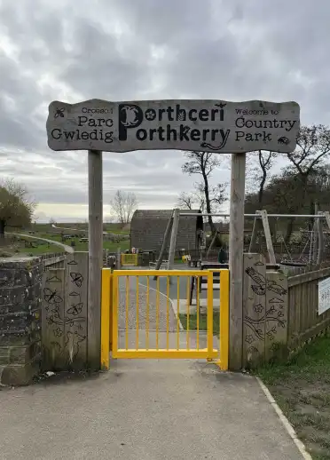 Wooden entrance sign and yellow gate welcoming visitors to Parc Gwledig Porthceri, with a playground and park paths beyond.
