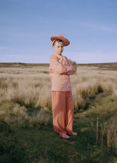 A child wearing a pale pink outfit and matching hat stands in an open grassy landscape under a wide blue sky. The person faces the camera with their arms crossed in a peaceful outdoor setting.
