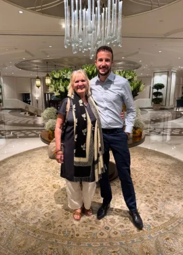 Two people smiling for a photo in an elegant hotel lobby, standing beneath a large glass chandelier with floral arrangements and decorative furnishings in the background.