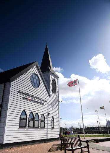 Imagen exterior de edificio blanco con campanario y asta de bandera con una bandera noruega a la derecha del edificio