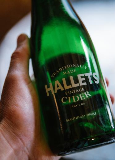 Une main tenant une bouteille verte labellisée Hallets Cider.