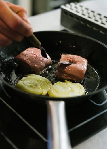 A chef's hand cooks meat in a frying pan on a stove in a kitchen.
