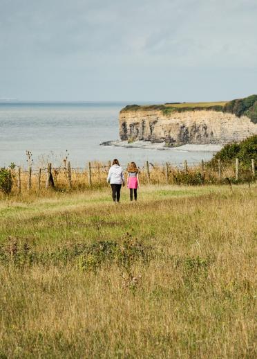 Two women walking along the coast, with high cliffs and a lighthouse in the background.