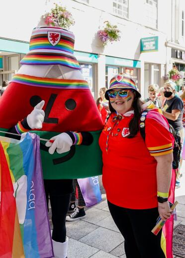 Tracy Brown at the Pride Cymru march wearing the Wales strip standing next to the Urdd mascot 