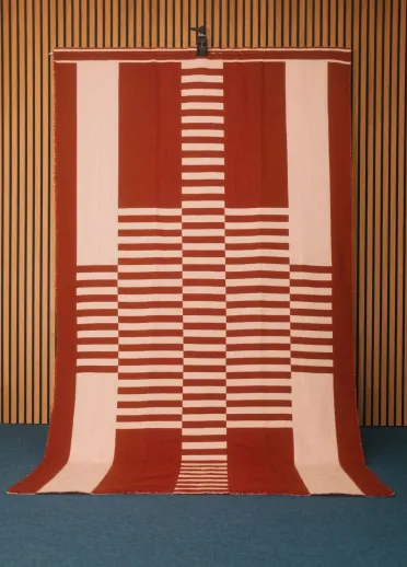 Una gran manta de rayas rojas y blancas, tejida a mano por la artista textil galesa Llío James. La manta está colgada en una pared de madera.