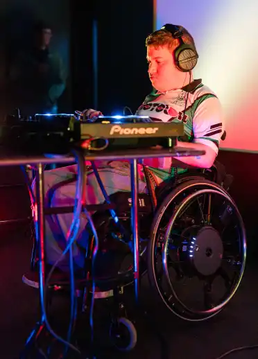 A person in a wheelchair using DJ equipment under colorful stage lighting. They appear focused while performing.