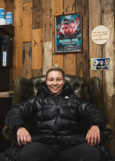 Lauren Price MBE sits smiling in an armchair inside a local barbershop, with boxing memorabilia and a promotional fight poster displayed on the wooden wall behind her. 