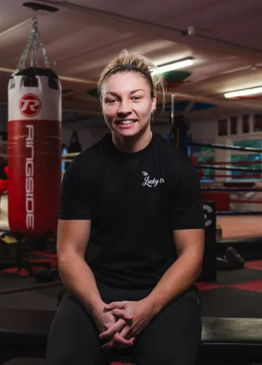 Lauren Price MBE sits smiling on a bench in a boxing gym, with a boxing ring and hanging punch bags visible in the background. 