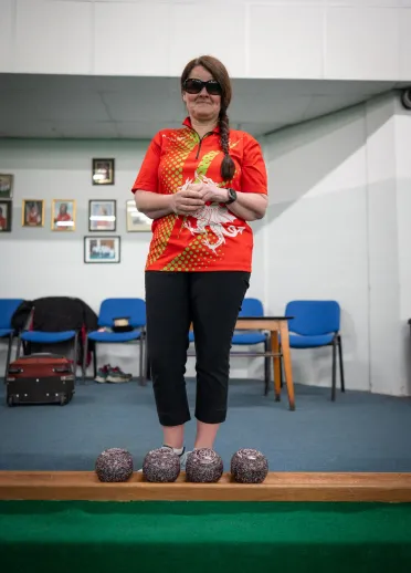 Julie Thomas standing with her bowls on an indoor bowling green in Team Wales kit.