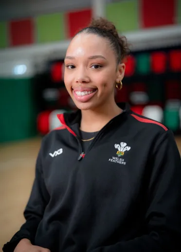 Portrait of Team Wales netball player Nansi Kuti in Welsh Feathers training kit, smiling at an indoor training venue.