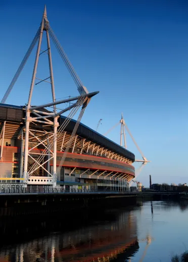 Exterior of the Principality Stadium in Cardiff, showing its distinctive steel masts and cables reflected in the River Taff beneath a clear blue sky. 