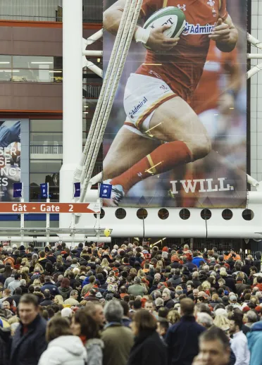 Crowds of rugby supporters gathering outside the Principality Stadium in Cardiff, beneath a large Welsh rugby banner. 