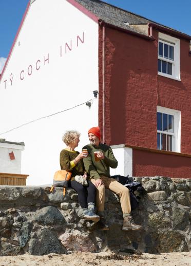 pareja sentada bebiendo en una pared mirando al mar fuera de Ty Coch Inn Porthdinllaen