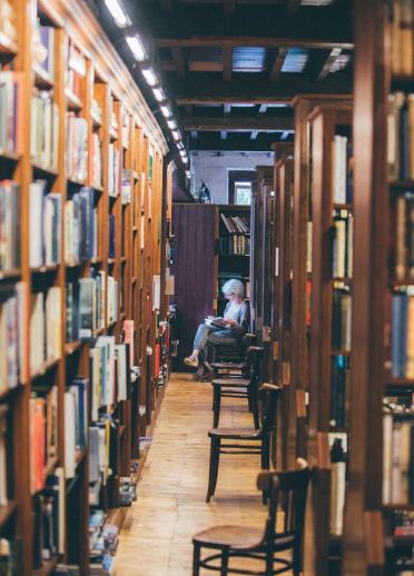 Eine Dame sitzt und liest in einer Bibliothek, umgeben von hohen Regalen voller Bücher.
