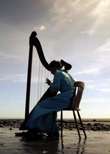 A photograph in silhouette of a young person wearing a blue dress playing a harp on a beach
