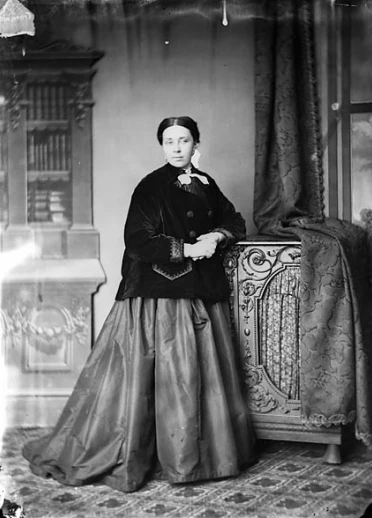 Dressed in a black button up jacket, a poufy long skirt and white accessories as she gazes to the right of the photo
