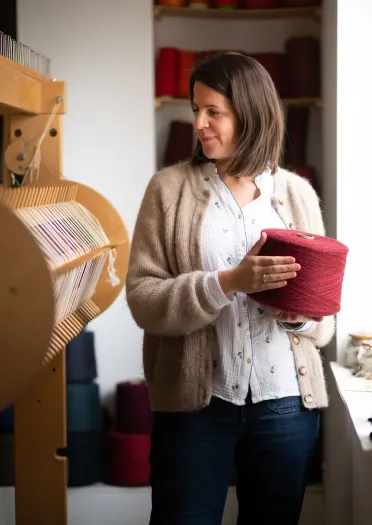 Welsh textile artist Llio James in her studio, holding a reel of red wool and looking at her hand loom