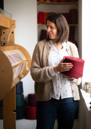 La artista textil galesa Llío James en su estudio, sosteniendo un carrete de lana roja y mirando su telar manual.