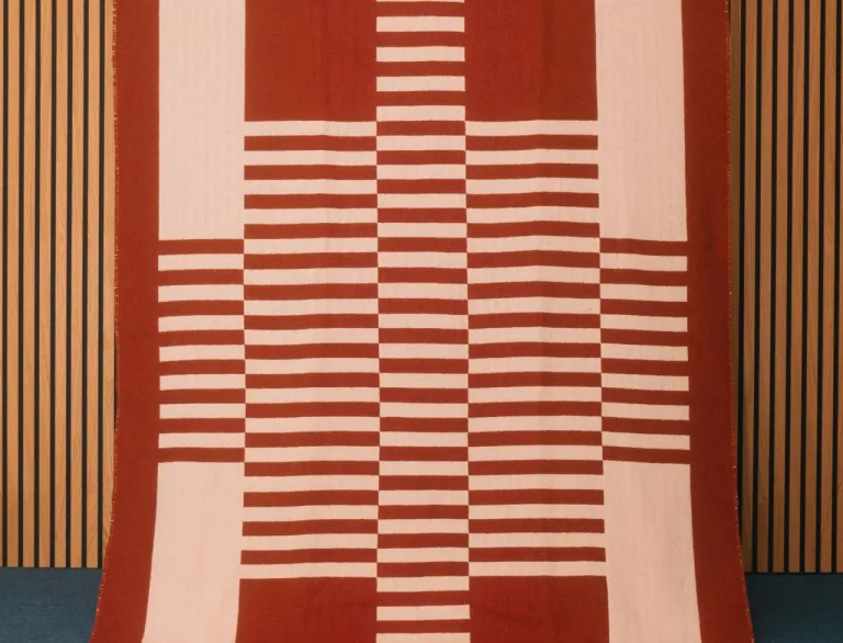 A large hand-woven red and white blanket in a striped pattern created by Welsh textile artist Llio James. The blanket is hanging on a wooden wall