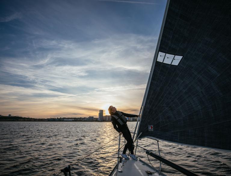 Hannah Mills leaning back from Main sail on yacht with sun setting on Cardiff Bay