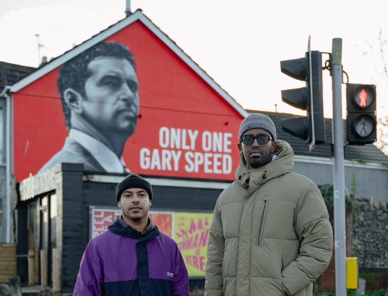 Two men standing in front of a mural of a man on the side of the building. The text reads 'Only one Gary Speed'.