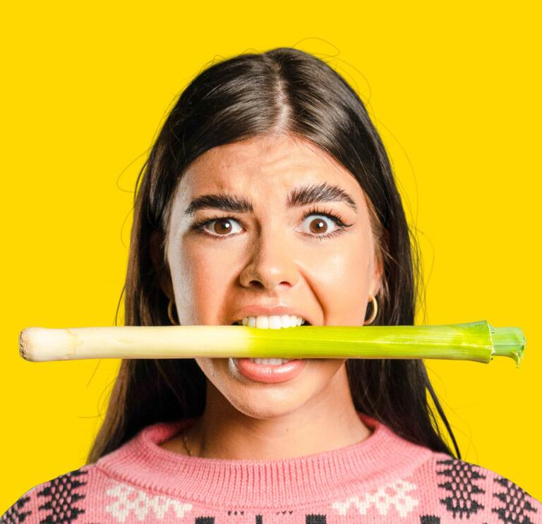 A woman wearing a pink jumper and holding a leek in her mouth