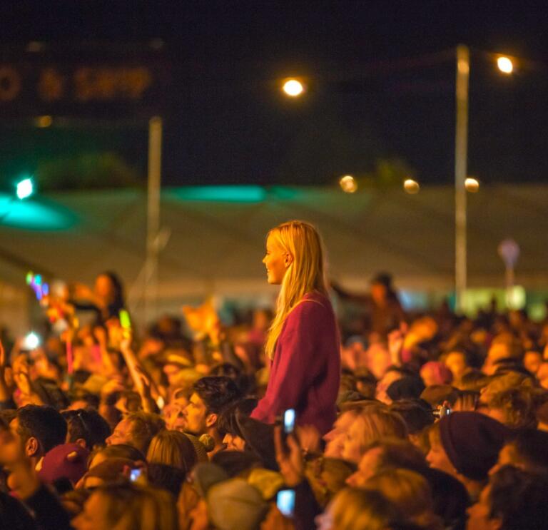 A crowd scene at a festival, with a girl sitting on someone's shoulders looking towards a stage