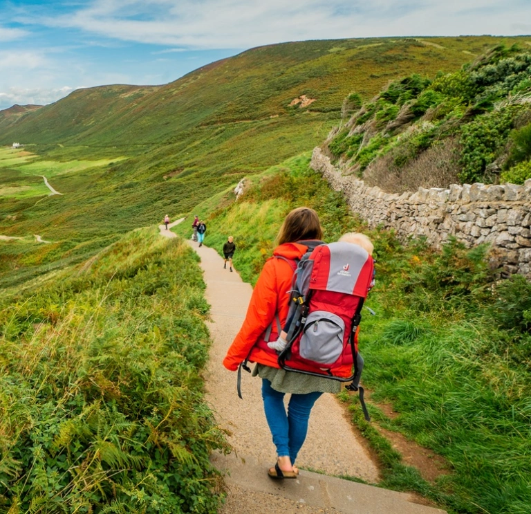 A parent with a baby in a hiking carrier walking down a beautiful coastal path with green to the right and the beach and blue sea to the left  