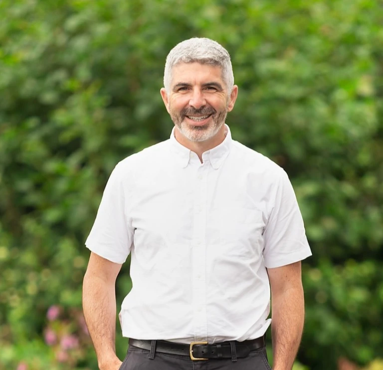 A man in a white shirt standing in front of green trees.