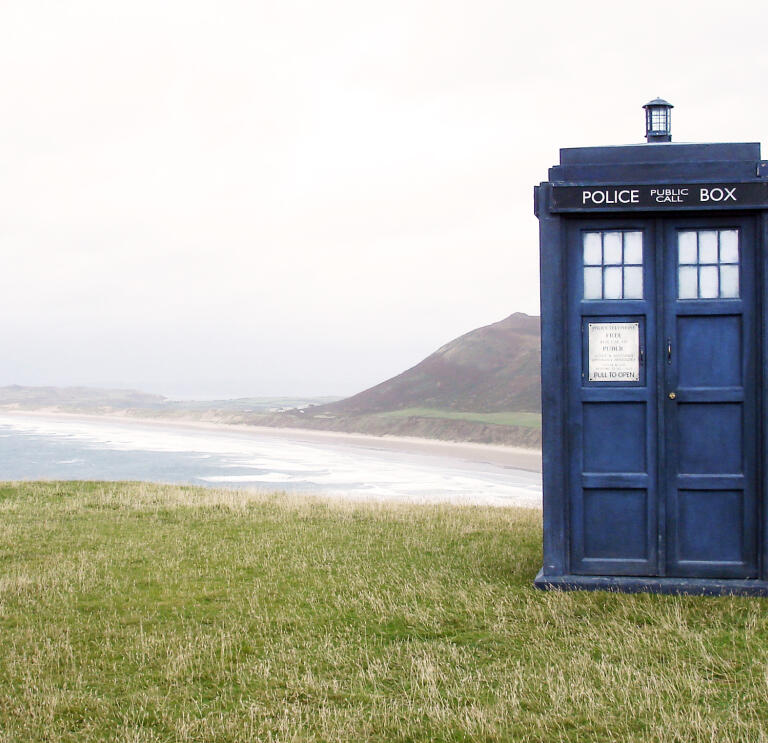 A blue phone box sat on grass with the sea in background.