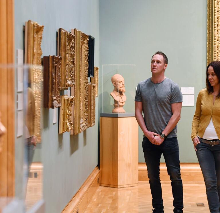 Two people walking through a gallery looking at paintings on the walls.