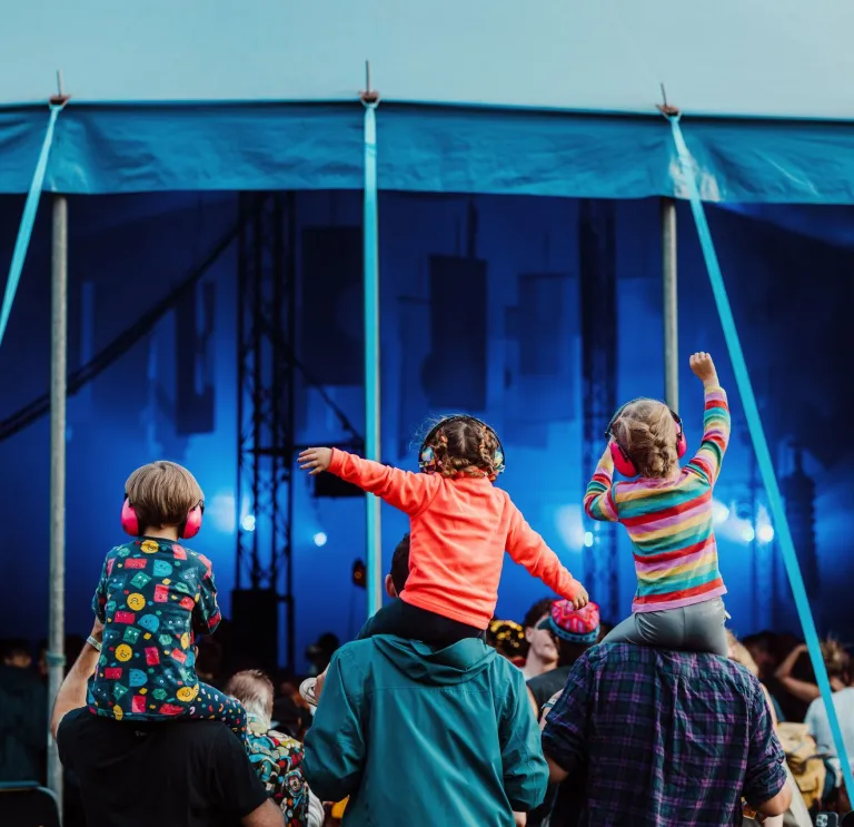 Three children sat on the shoulders of their parents listening to a band playing at a festival.
