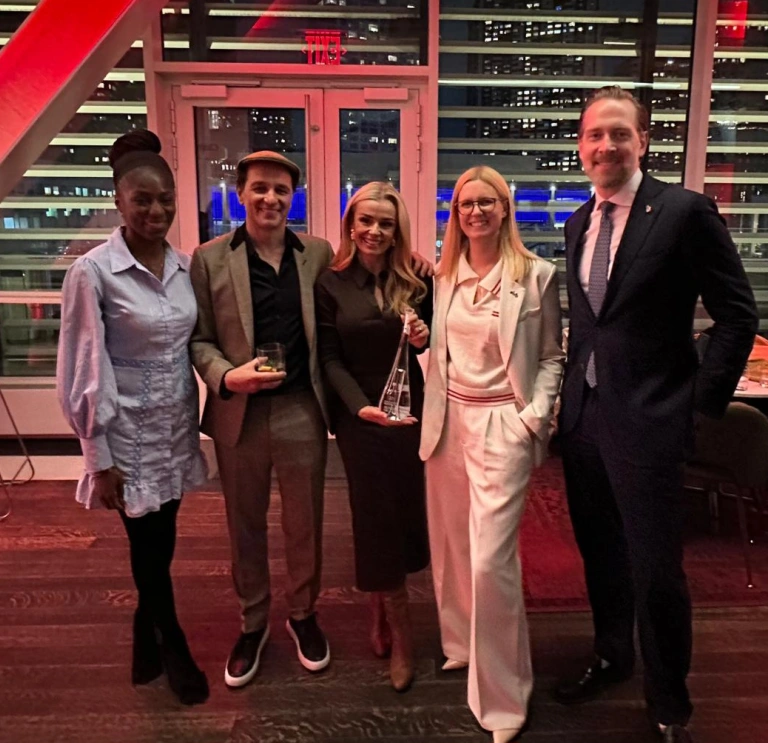 A group of five people pose together at an indoor event with city lights visible through the windows.