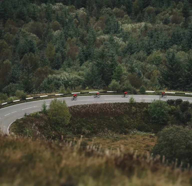 Four cyclists in red jerseys cycling round a tight bend in a mountainous road, surrounded by a mixture of beautiful trees