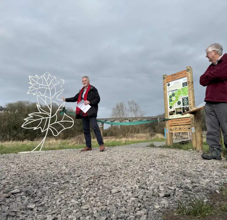 A man speaks beside a leaf shaped metal sculpture and a green ribbon across a gravel path at a country park entrance, with others watching and a viaduct in the background.