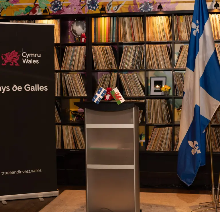 Podium with small Welsh and Québec flags, beside a Cymru Wales “Pays de Galles” banner, with full sized Québec and Wales flags and shelves of vinyl records behind.