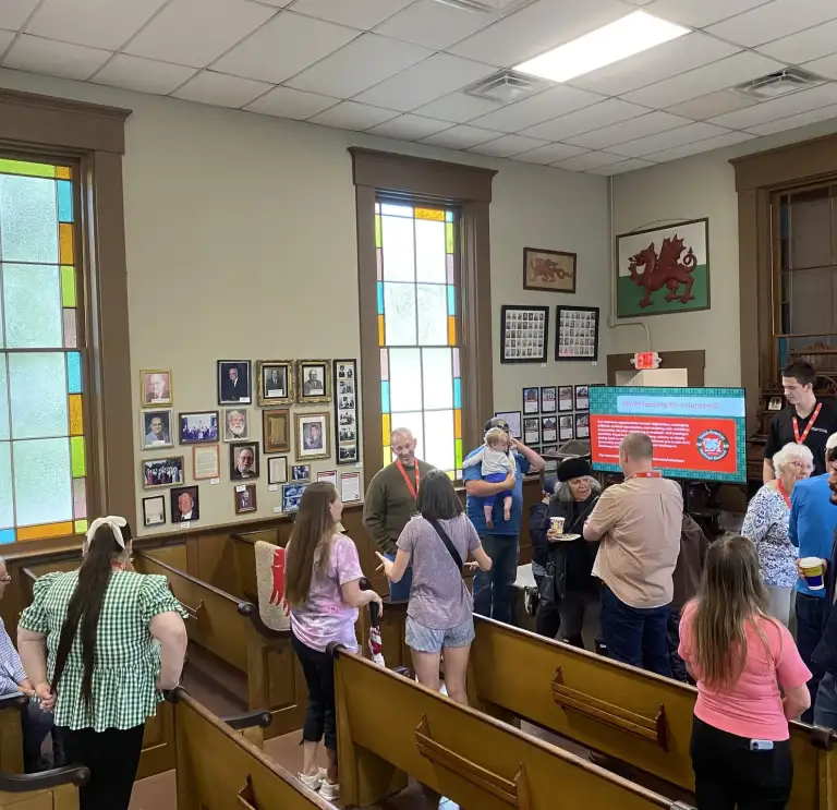 People gather and talk inside a historic building. A presentation screen and colorful windows are visible in the background. 