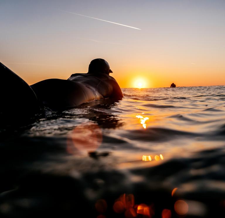 Surfer in wetsuit floating on the surface of the sea, silhouetted by the sunset on the horizon