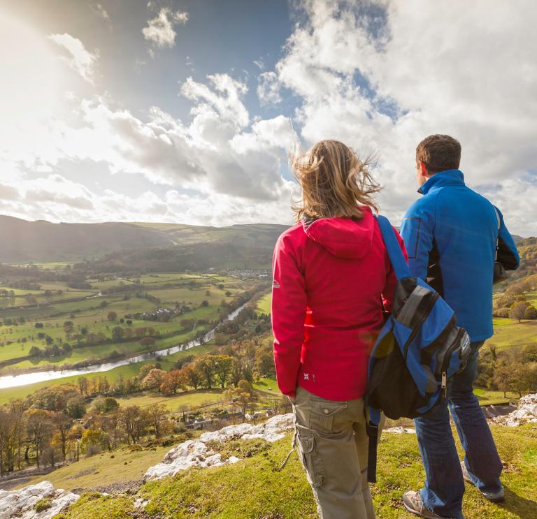 female and male walkers in the foreground looking down on a green valley  with a river