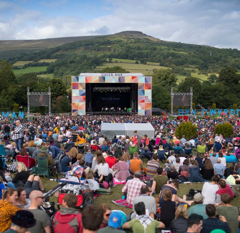A crowd of people sat in a field looking towards an outdoor festival stage with a hill in the background