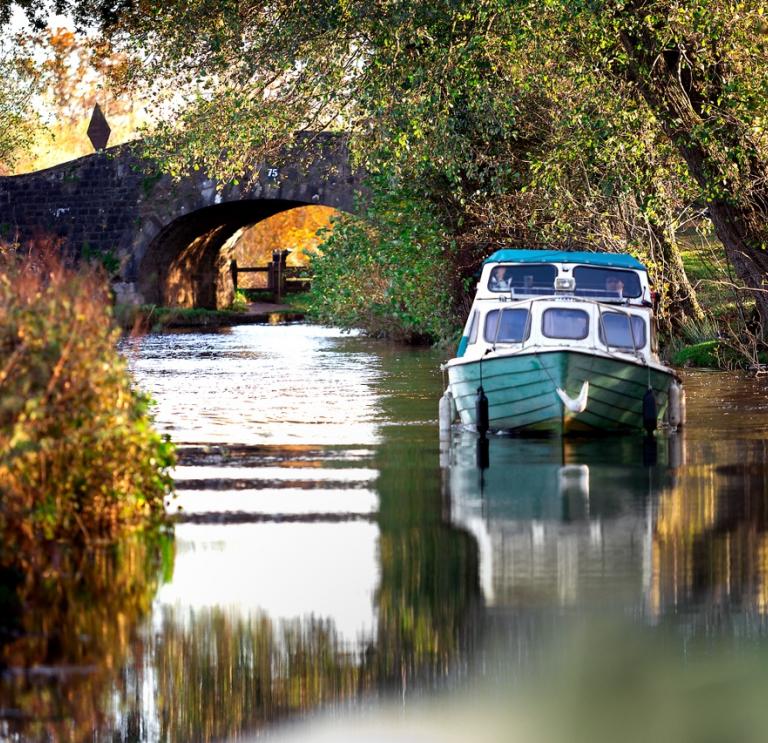 Hausboot auf einem Kanal umgeben von Bäumen, mit einer Brücke aus Stein im Hintergrund.