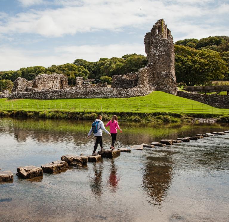 Horses and walkers in front of a ruined castle surrounded by green fields and water.