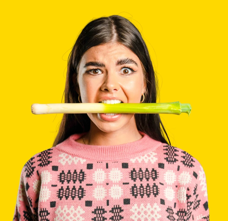 Fille portant un pull traditionnel gallois en maille, avec un poireau dans la bouche et une expression confuse sur le visage