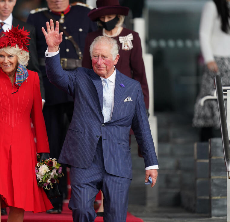 King Charles III accompanied by the Queen Consort, walking down some stairs and waving at crowds
