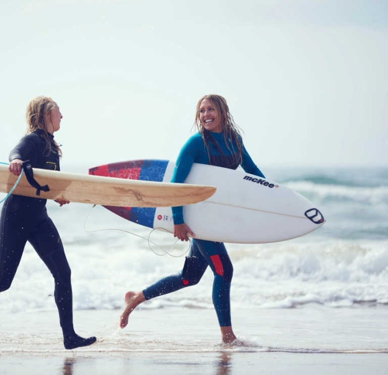 Dos personas con trajes de neopreno y sosteniendo tablas de surf corren a través del agua sobre la arena en una playa.