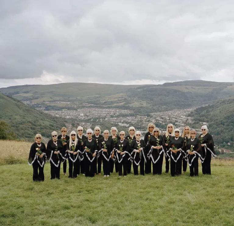 El coro femenino Dare to Sing, vestido de negro, de pie sobre un prado verde frente a los valles ondulados de Gales.