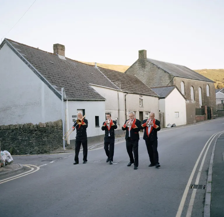 Cuatro miembros de una banda de metales tradicional galesa tocando sus instrumentos, caminando por una calle típica de los valles galeses.