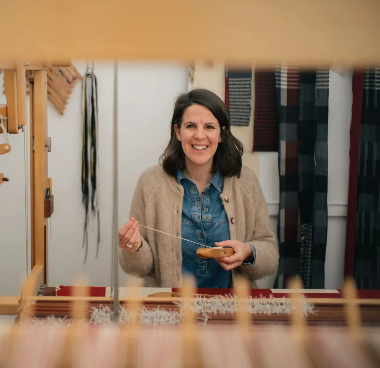 La artista textil galesa Llío James sentada en su telar manual, sonriendo a la cámara.