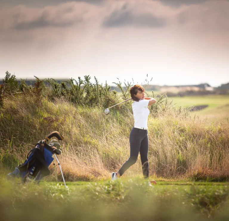 Person playing golf on an open course under a cloudy sky, captured mid-swing with a golf club and golf bag beside them.