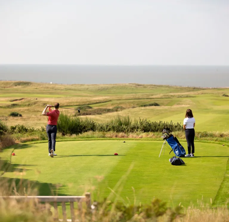 Scenic coastal golf course with one golfer preparing to swing and another observing with a golf bag.